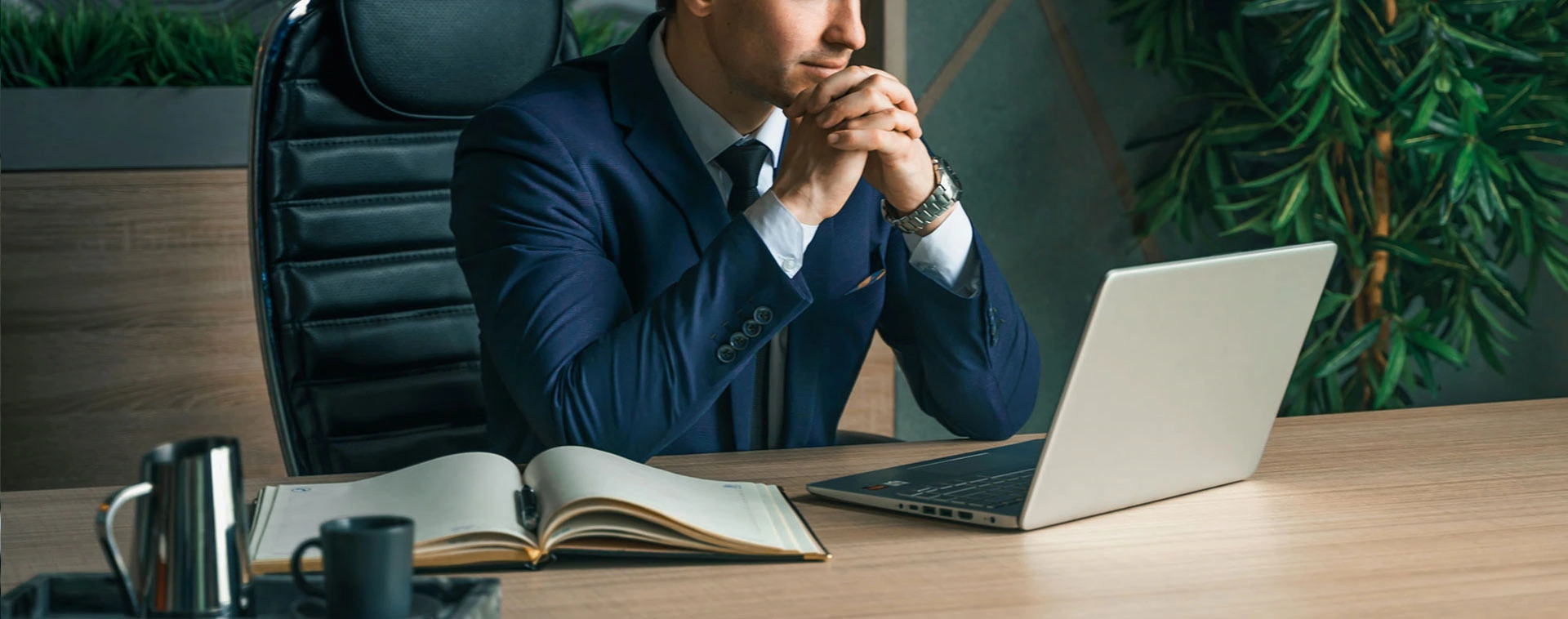 A man in a suit sits at a desk with a laptop, an open notebook, and a pen holder, appearing to think while reviewing mergers and acquisitions advisory documents on the screen.